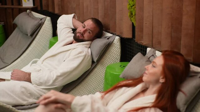High-angle view of young loving husband and wife in white bathrobes conversating while lying hand in hand on loungers near indoor pool in luxury spa hotel. Happy couple spending vacation together.