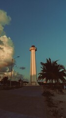 Fotografia de un faro con la mar y precencia de palmeras marcadas por las sombras al costado derecho