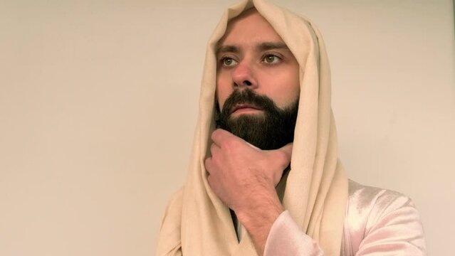 close-up male face in beige head cape, young pensive bearded man in image of Jesus Christ, satisfied guy with 30 years seriously looks away, concept Christian religion, holiday of Easter, Christmas