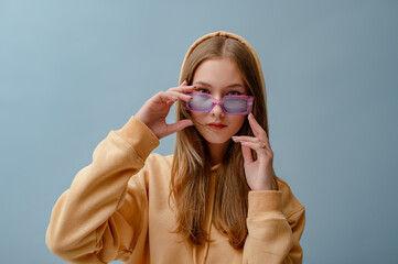 Fashionable confident girl wearing purple sunglasses, yellow hoodie,  posing on blue background....