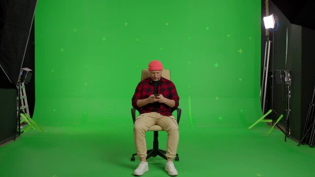 Full body video shot of man in casual clothes sitting in office chair texting messages holding phone on green screen background