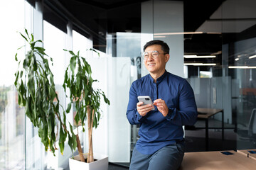 Successful asian man inside office looking out the window, boss in shirt holding smartphone, man in...