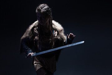 A young brave Viking heroine with a sword in hands on a dark background. A young woman in a suit in...
