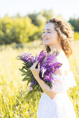 Fototapeta premium Beautiful young girl in a white dress, straw hat, with picnic and bouquet of purple wild flowers on a meadow. Summertime, golden hour, sunset gathering plants for handmade organic cosmetic production