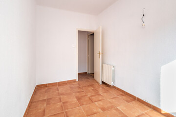 An empty spacious room with old square worn caramel colored floor tiles and a radiator on the wall, with an open door, white faded walls and ceiling. The wires for the lamp stick out in the wall.