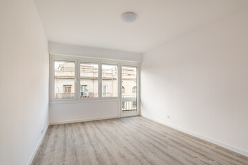 An empty bright room with gray wood parquet, white walls visually make the room even larger. Room has windows and a door with access to a small balcony and a view of the house opposite.