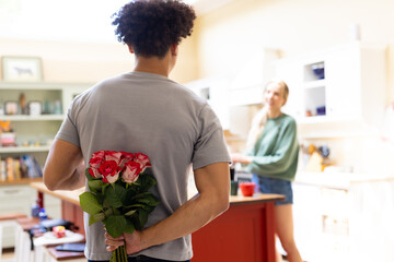 Rear view of biracial young man hiding red roses behind back while talking with wife at home