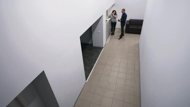Worker stands on reception with female secretary, meets and greets walking coworker at hall. Men discussing plans of project at corridor. Lobby at business center of office. Security camera shooting.