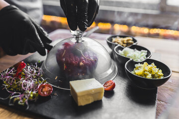 Unrecognisable person lifting transparent lid off of elaborate smoked beef tartare served on black...