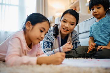 Happy Chinese mother talks to her kids while drawing on paper together at home.