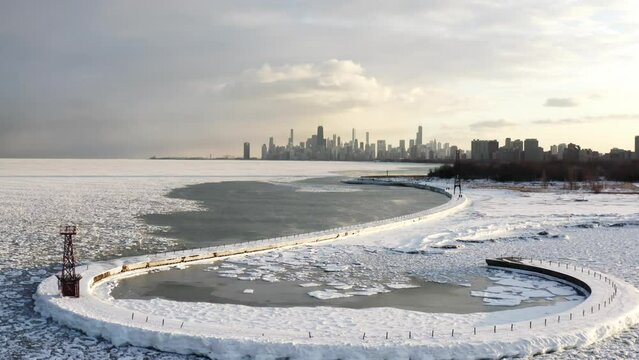 Panning in winter aerial cityscape scene flying towards a hooked shaped pier with snow encrusted ice chunks floating on Lake Michigan below and Chicago skyline in the distance at sunset. 