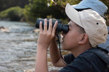 Asian boys wearing black t-shirt holding a binoculars sitting on stone by the river flowing down from the mountains in national park to observe fish in the river and birds on tree branches and on sky.