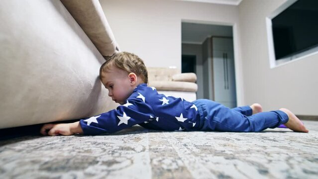 Toddler Boy Lies On The Floor Reaching His Hand Under The Sofa. Baby Wants To Get Something From Under The Couch.