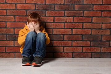 Upset boy sitting on floor near brick wall, space for text. Children's bullying - obrazy, fototapety, plakaty