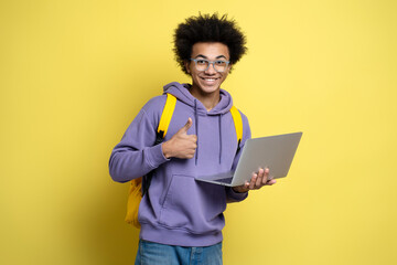 Attractive smiling African American man freelancer using laptop holding thumb up isolated on yellow background. Portrait of smart happy student studying, education concept