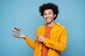 Young smiling African American man holding shopping trolley with credit card looking at camera isolated on blue background. Shopping, sales concept