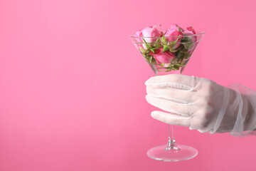 Woman in elegant gloves holding beautiful martini glass with water and roses on pink background,...