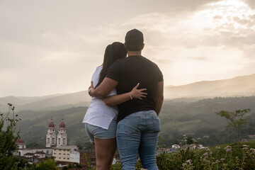 Young couple embracing outdoors and contemplating the natural landscape and an old Latin American colonial town. Appreciating the sunset in the mountains - Charalá, Santader, Colombia