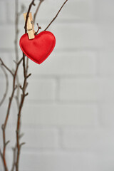 A red cloth heart hangs on a clothespin on the branches of a tree against a background of white brick. Vertical location. The concept of Valentine's Day.