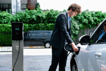Progressive businessman with electric car recharging at public charging station at modern city...