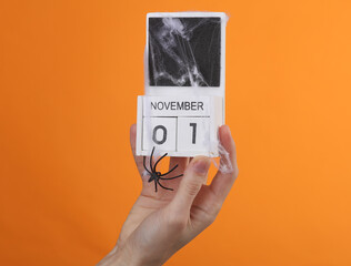 Happy Halloween. Hand holding wooden calendar with date November 01 and halloween decor on orange background