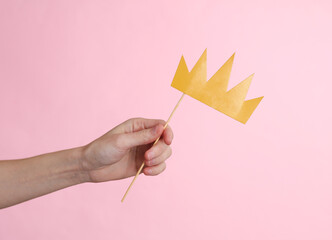 Hand holds paper cut golden crown with stick on pink background. Party concept
