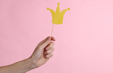 Hand holds paper cut golden crown with stick on pink background. Party concept