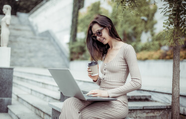 Young stylish woman in sunglasses uses a laptop while sitting on a bench with a coffee in a city park