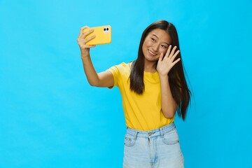 Asian woman holding her phone and looking at the screen taking pictures of herself talking on a video call with a yellow case on a blue background wearing a yellow T-shirt smiling with teeth 