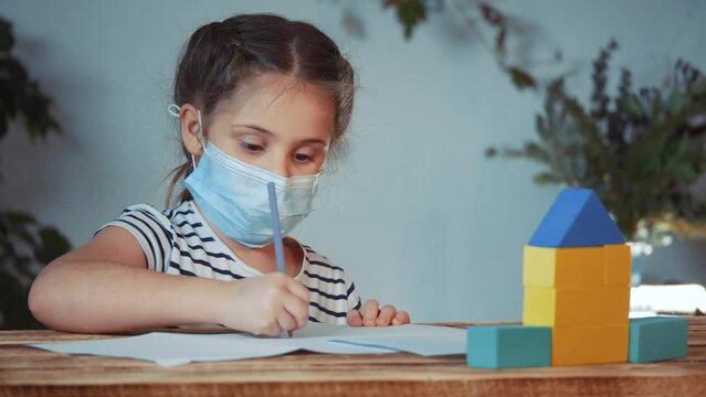 little girl painting at home wearing protective medical mask. coronavirus learning at home remotely kid a dream concept. girl kid studies remotely in a protective medical mask. school fun home dream
