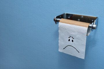 Sad face on the last piece of toilet paper, tissue on a brown cardboard toilet roll, blue wall background, copy space