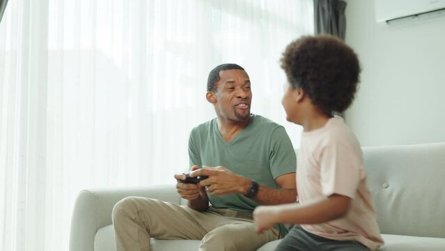 Black African American Father and his little son sitting on the couch playing video games at home, Having fun.
