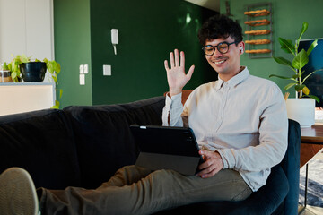 Young multiracial man smiling while using digital tablet and waving on sofa with feet up at home