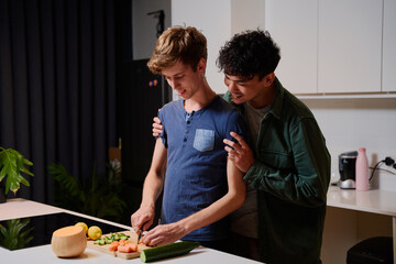 Young gay couple in casual clothing smiling while preparing dinner in kitchen at home