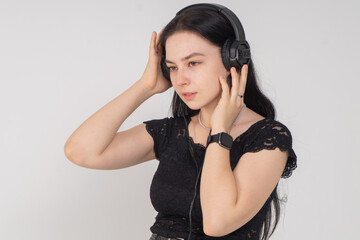 Attractive young brunette girl listens to music in large overhead headphones. A music lover is dressed in a black lace blouse and trousers, she is posing on a white background