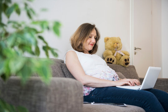 Pregnant woman sitting on sofa and working on laptop, Munich, Bavaria, Germany