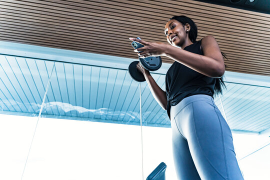 Young black girl in the gym doing exercises with a barbell and phone