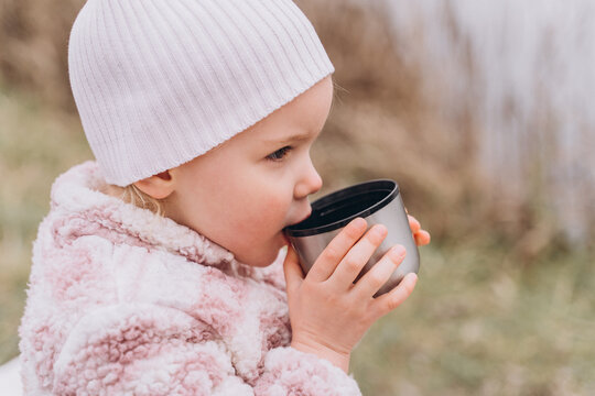 Baby girl in light pink warm clothes drinks tea from a thermos
