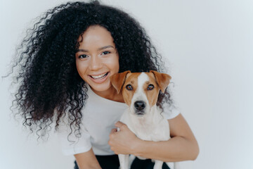 Close up shot of beautiful happy Afro woman with bushy curly hair, embraces favourite dog and have...