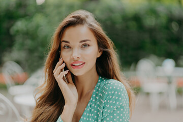 Fototapeta premium Horizontal shot of pretty young woman wears polkadot blouse, makes phone call, speaks with friend, poses outside against green blurred background, wears lipstick. People and communication concept