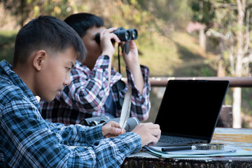 Naklejka premium Asian boys are at the local national park, they are consulting, talking, doing their school project work as bird watching topic by using binoculars, laptop, map and example bird feathers to compare.