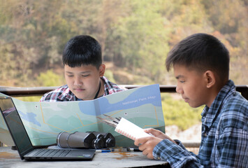 Asian boys are at the local national park, they are consulting, talking, doing their school project work as bird watching topic by using binoculars, laptop, map and example bird feathers to compare.