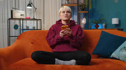 Obraz premium Woman with short blonde hair sitting on sofa uses mobile phone smiles at night home apartment. Girl texting share messages content on smartphone social media applications online, watching relax movie