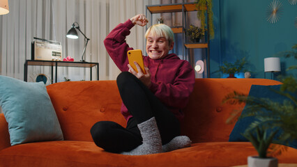 Obraz premium Happy woman with short hair use mobile smartphone typing browsing say Wow yes found out great big win good news celebrate lottery jackpot doing winner gesture. Girl at home in room sitting on sofa