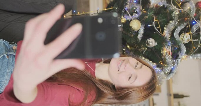 Girl takes selfie photo on smartphone, of herself with glass champagne against background of Christmas tree, standing at home in the living room. Vertical video.