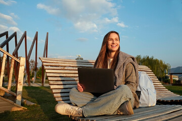 Young smiling woman sitting sitting with the laptop and working on the wooden deck chair in the park. Natural soft sunset light, copy space, background.