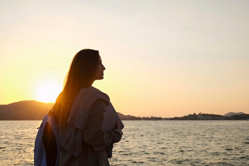 Young woman wearing smart casual outfit standing by the sea with laptop in her hands and enjoying the scene of sun setting over the islands. Copy space, background.