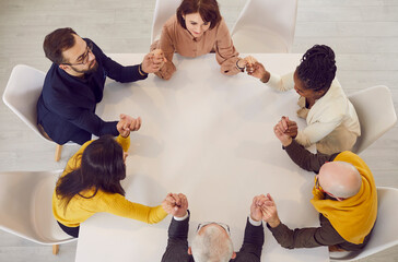 Team of business people sitting around table holding hands. View from above of male and female...