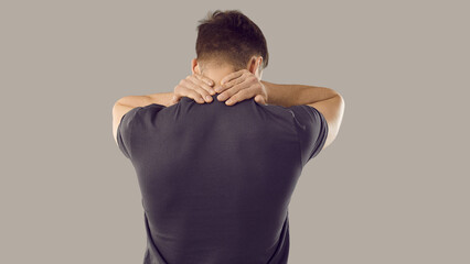 Rear view of tired young man rubbing his sore neck, isolated on gray background. Unknown muscular...
