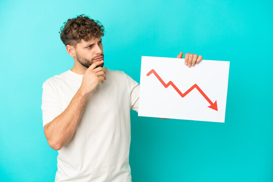 Young handsome caucasian man isolated on blue background holding a sign with a decreasing statistics arrow symbol and thinking